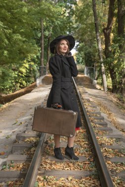 Geniş kenarlı şapkalı, siyah ceketli ve eski moda valizli akıllı bir kadın parkta yürüyor. Dikey çerçeve.