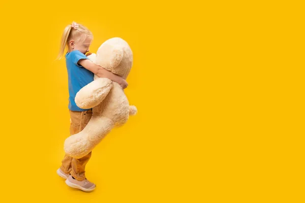 Girl of 5-6 years old hugs huge teddy bear. Full-length portrait of preschool child with toy in studio on yellow background. Copy space.