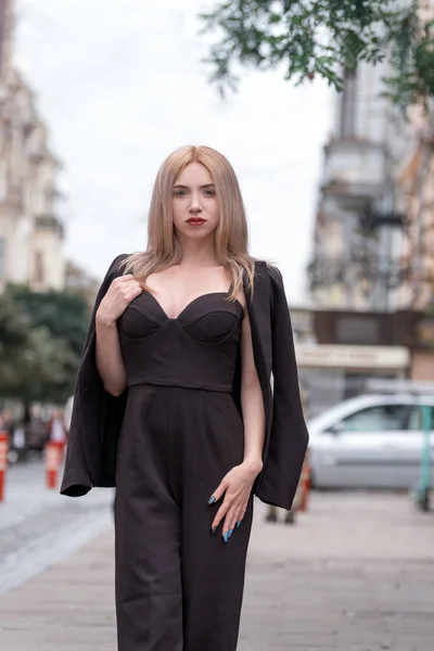 ビジネススーツのエレガントな女性が街を歩き回っています 若い女は深いネックラインと黒のスーツでブロンドです — ストック写真