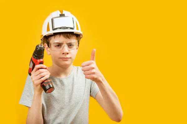 十几岁的男孩戴着防护头盔和眼镜 拿着钻头 把大拇指伸出来 与黄色背景隔离 作为建设者的孩子复制空间 — 图库照片