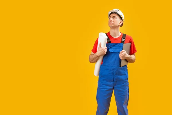 身穿蓝色工作服的建筑工人或人工工程师戴着安全帽 拿着图纸和文件抬头看 被黄色背景隔离 复制空间 — 图库照片