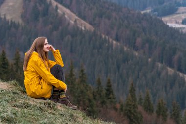 Orman arka planında, yamaçta uzun saçlı güzel bir genç kadın. Ormanda sarı yağmurluk giyen güzel kız..