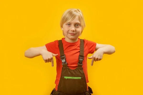 一个金发碧眼的小学生穿着红色T恤和连衣裙的画像 他的食指朝下 对着相机笑着 黄色背景 — 图库照片