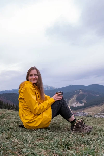 Genç bir kadın yamaçta oturur, çay içer ve gülümser. Dağların arkasındaki sarı parktaki turist kızın portresi. Dikey çerçeve