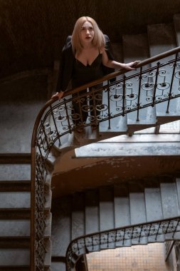 Koyu yakalı siyah elbiseli güzel sarışın kız eski bir merdivende duruyor. Üst Manzara. Dikey çerçeve