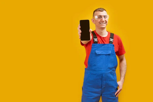 身穿制服和护目镜的工人微笑着 手持带有空白黑屏的智能手机 在黄色背景下被隔离 复制空间 — 图库照片