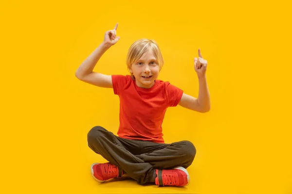 一个十几岁的男孩 一头金发 双腿交叉地坐着 用食指尖着他的食指 在黄色背景下隔离 — 图库照片