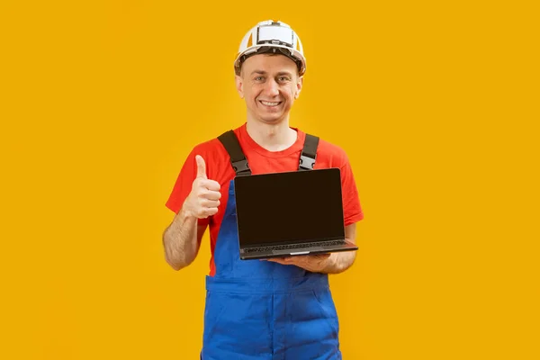 身穿蓝色套头衫 头戴安全帽的男人拿着带有空白黑色屏幕的笔记本电脑 竖起大拇指 摆出一副样子 复制空间 — 图库照片