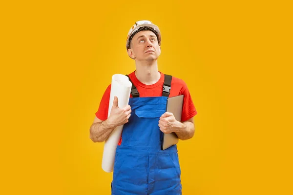 身穿蓝色工作服的土木工程师或建筑工人戴着安全帽 拿着图纸和文件抬头看 在黄色背景下隔离 — 图库照片