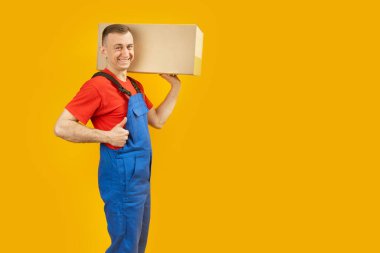 Sarı arka planda karton kutu tutan sıradan bir genç adam baş parmağını kaldırıyor. Sarı arka planda izole edilmiş. Uzayı kopyala, taklit et