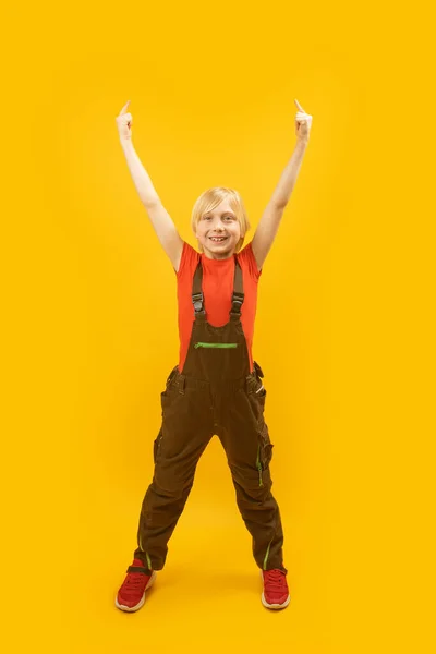 快乐的金发男孩举起双手 手指指向某个地方 穿着黄色背景工作服的青少年的画像 复制空间 — 图库照片