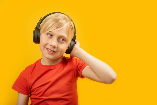 ブロンドの少年は赤いTシャツを着て音楽を聴く 黄色の背景に隔離されたヘッドフォンのティーンエイジャーの肖像画 スペースのコピー — ストック写真