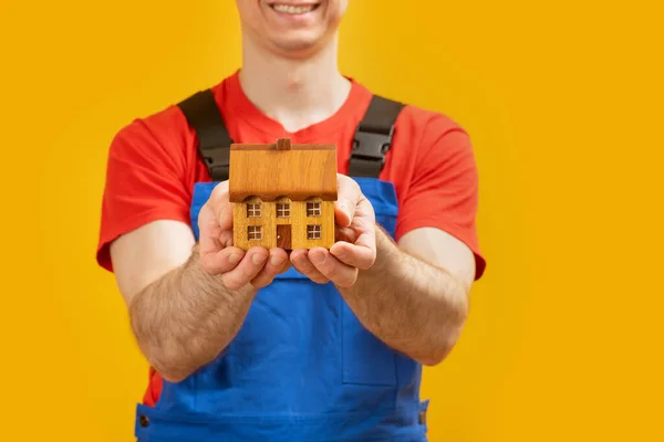 Man in worker uniform holds model at home. Small toy wooden house in mens hands. Close up.
