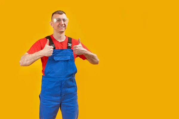 身穿蓝色制服 面带微笑的年轻工人或建筑工人被隔离在黄色的背景下 露出大拇指向上看 复制空间 — 图库照片