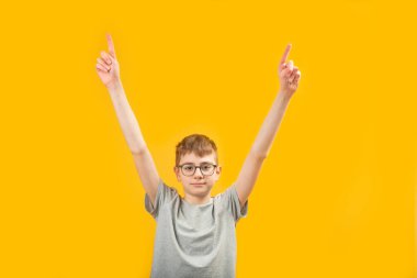 Beyaz tenli esmer çocuk gözlük takıyor ve gri tişört giyip işaret parmaklarıyla ellerini kaldırdı. Sarı arka planda izole edilmiş. Boşluğu kopyala