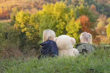 Kardeşler sonbahar gününde. Sarışın erkek ve kız kardeş açık alanda büyük bir oyuncak ayıyla oturuyorlar. Arka plan