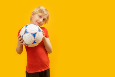 Kırmızı tişörtlü sarışın çocuk futbol topu tutuyor. Sarı arka planda topu olan genç kızın stüdyo portresi. Boşluğu kopyala