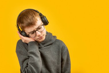 Boy with glasses and hoodie listening to music with headphones. Teenager wears headphones listens to audiobooks, podcasts or lessons. Copy space clipart