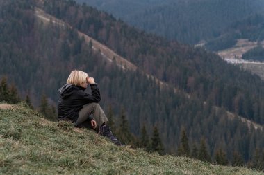 Sarışın çocuk dağın yamacında oturur ve uzaklığa bakar. Çocuk rahat. Çocuklarla dağlarda hafta sonu