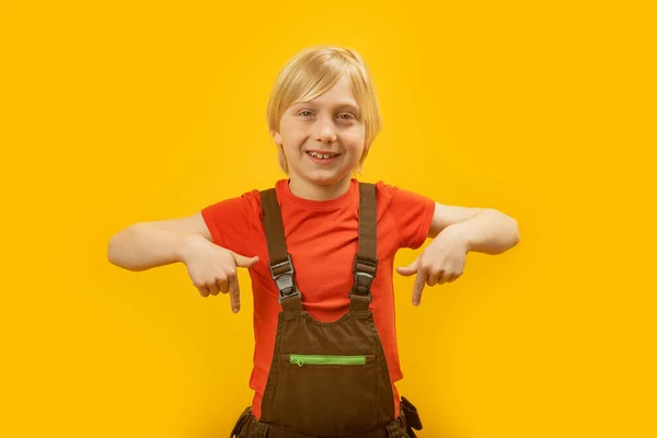 赤いTシャツを着た金髪の少年の肖像画と 人差し指を下にしてカメラに向かってウィンクするジャンプスーツ 黄色の背景 — ストック写真