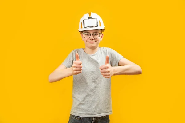 戴眼镜的男孩戴着防护头盔 用双手竖起大拇指 在黄色的背景上 青少年们摆出一副姿势 — 图库照片