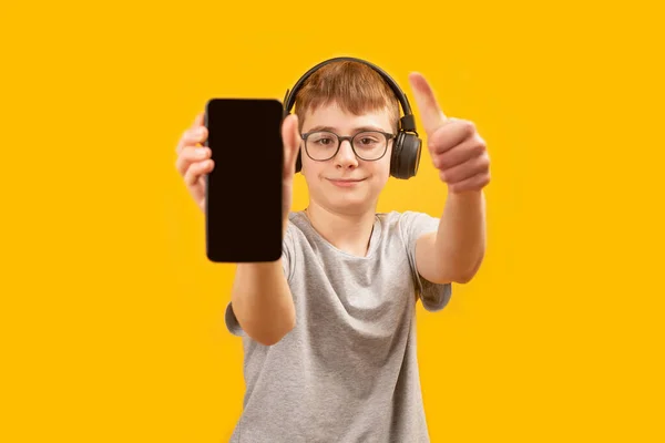 ヘッドフォンやメガネの学校の少年は 空白の画面でスマートフォンを保持し 親指が表示されます 音楽や学習のためのアプリ スペースのコピー — ストック写真
