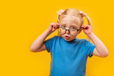 4 yaşındaki şaşırmış bir kız gözlüğünü ayarlıyor. Sarı arka planda gözlüklü mavi tişörtlü küçük sarışın kızın portresi.