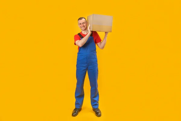 Sarı Arka Planda Omzunda Bir Kutuyla Mavi Tulumlu Bir Erkek — Stok fotoğraf