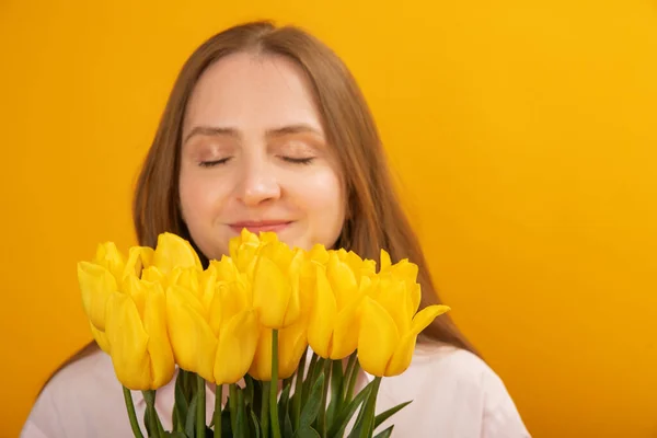 stock image Young blonde woman enjoys scent of flowers. Girl holds bouquet of yellow tulip flowers on yellow background. Close up. Copy space