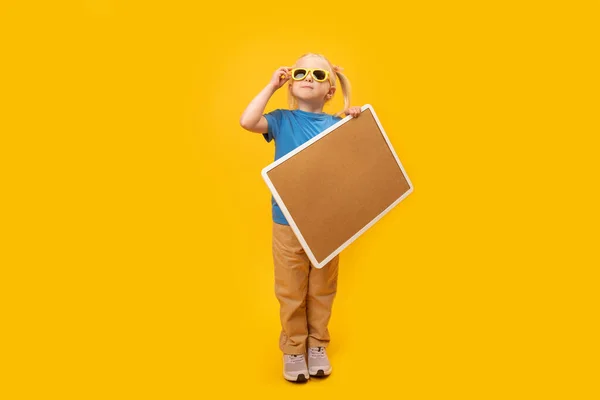 stock image Little blonde girl with two ponytails wears sunglasses and holds cork board. Portrait of child with blank board on yellow background