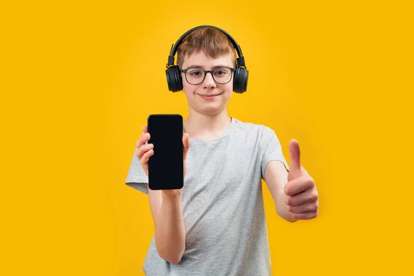 メガネとヘッドフォンを身に着けている学校の少年は 空白の画面でスマートフォンを保持し 親指を表示します オーディオブックや音楽や学習を聞くためのアプリ コピースペース モックアップ — ストック写真