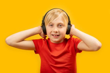 Kırmızı tişörtlü beyaz saçlı çocuk büyük siyah kulaklık takıyor ve kafasında tutuyor. Sarı arkaplanda izole edilmiş