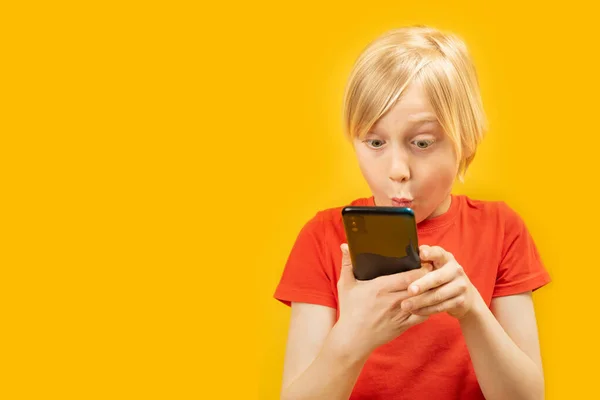 驚くべき10代の男の子がスマートフォンの画面を見ています 黄色の背景に彼の手に携帯電話で赤いTシャツの男性の子供の肖像画 スペースのコピー — ストック写真