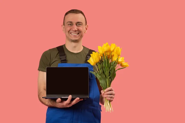 一个年轻男子的画像 他拿着一束鲜花和一个空的黑色屏幕的笔记本电脑 粉红的背景 复制空间 网上订购花卉 — 图库照片