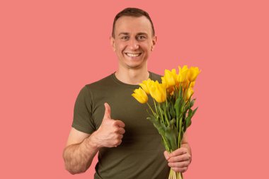 Yakışıklı, orta yaşlı, elinde bir buket sarı lale çiçeği tutan ve parmağını kaldıran bir adam. Pembe arka planda izole edilmiş stüdyo portresi