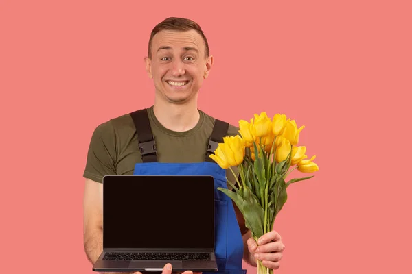Surprised young man with bouquet of flowers and laptop with empty black screen. Pink background. Order flowers online. Copy space, mock up.