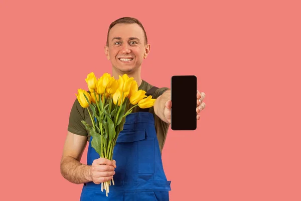 Portrait of happy flower delivery man holding smartphone with blank black screen isolated on pink background. Order flowers online in mobile application