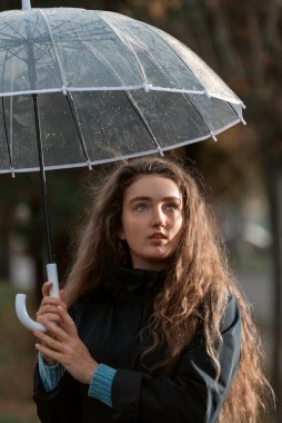 Dalgalı saçlı mavi gözlü kız, şeffaf şemsiyeyi elinde tutarken hüzünlü bir şekilde yan yana duruyor. Dikey çerçeve