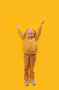 4-5 yaşlarında, at kuyruklu, küçük sarışın bir kız ellerini kaldırıp kameraya baktı. Sarı arka planda sarı elbiseli çocuğun stüdyo portresi