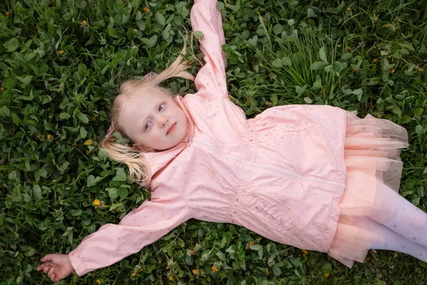 身穿粉色衣服和夹克的金发小女孩躺在绿草上 胳膊伸到两边 春天的一天顶部视图 — 图库照片