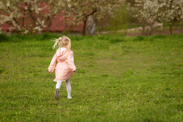 木の背景に対して緑色のメドウにピンクのジャケットで小さな金髪の少女を走らせます スプリングタイム 公園で楽しんでいる女の子 — ストック写真