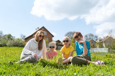 Evin arkasındaki çimlerin üzerinde oturan genç kadın ve üç çocuk. Evin arka bahçesinde aile pikniği