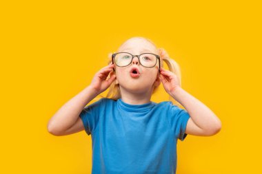 Şaşırmış küçük kız gözlüklerini ayarlıyor ve yukarı bakıyor. Sarı arka planda gözlüklü mavi tişörtlü küçük sarışın kızın portresi.