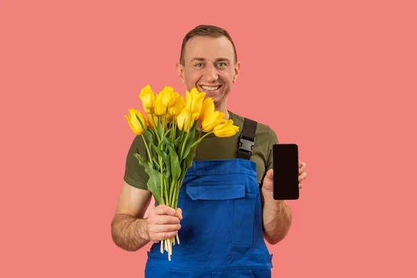Portrait of smiling flower delivery man holding smartphone with blank black screen on pink background. Order flowers online in mobile application