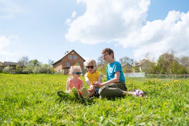 Çocuklar arka planda yeşil çimlere otururlar. Evin yakınındaki çayırda güneş gözlüklü üç çocuk.