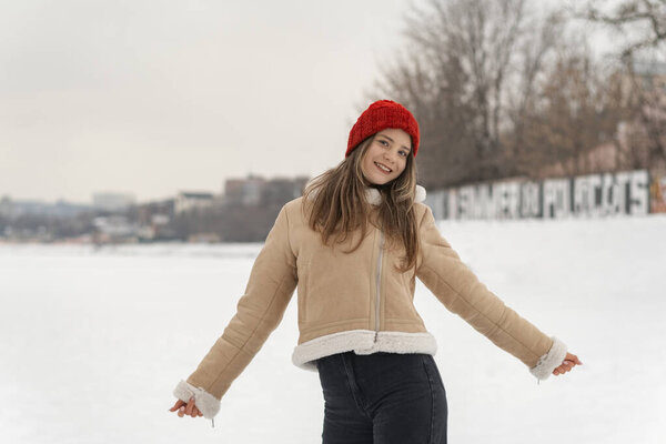 Portrait of joyful girl in red knitted hat and beige sheepskin coat with fur walking through snow-covered park.