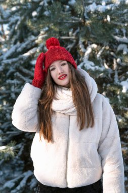 Kırmızı dudaklı güzel bir kadın karlı kış parkında şapkasını düzeltiyor, çam ağaçlarının arkasında. Güneşli kış günü.