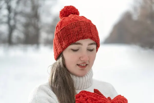 Young pretty woman warms up hands in woolen clothes outdoors, cold weather. Girl in knitted red hat and mittens, winter park.