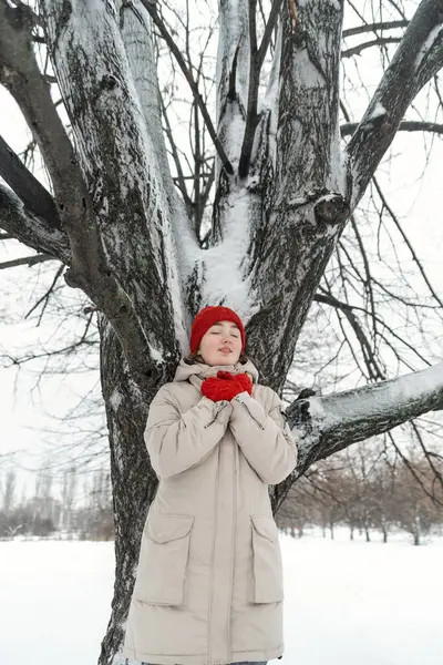 Beautiful girl in warm clothes stands with her eyes closed under tree in winter park. Woman takes pleasure being in nature.
