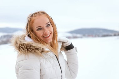 Beyaz ceketli, kızıl saçlı, gülümseyen ve kar yağışlı kış gününde kulaklıklı bir kadın.